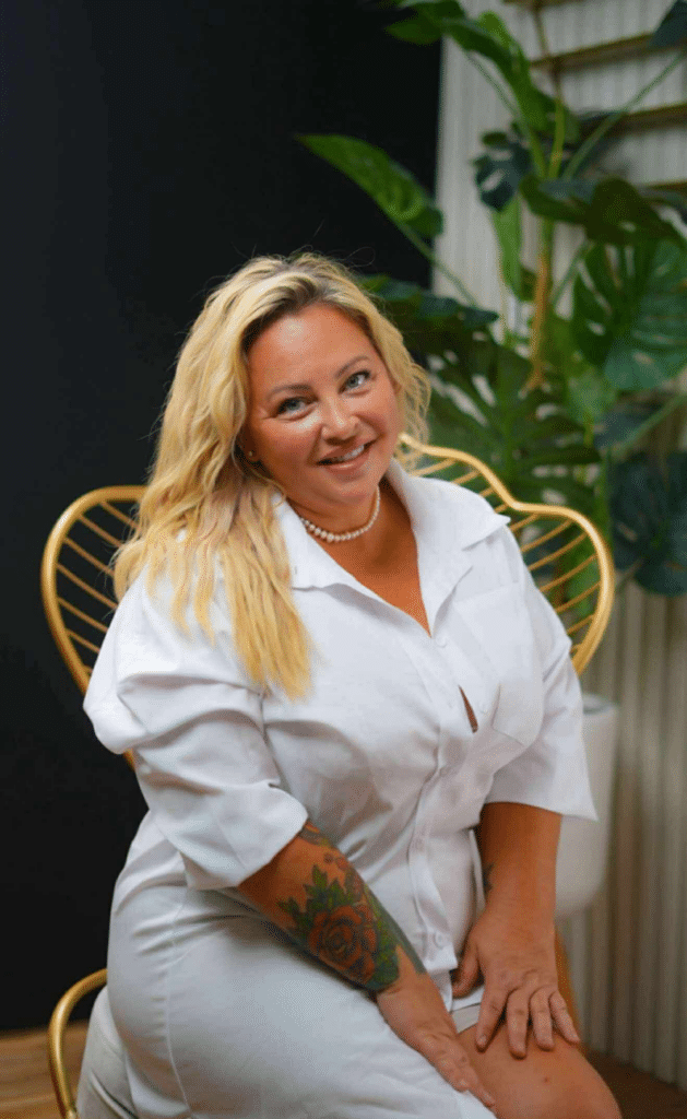 Smiling blonde woman in a white blouse sits on a gold, curved chair with indoor plants behind her.